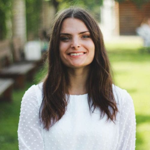 Smiling outdoor headshot of Kristina wearing a white top with greenery in the background.